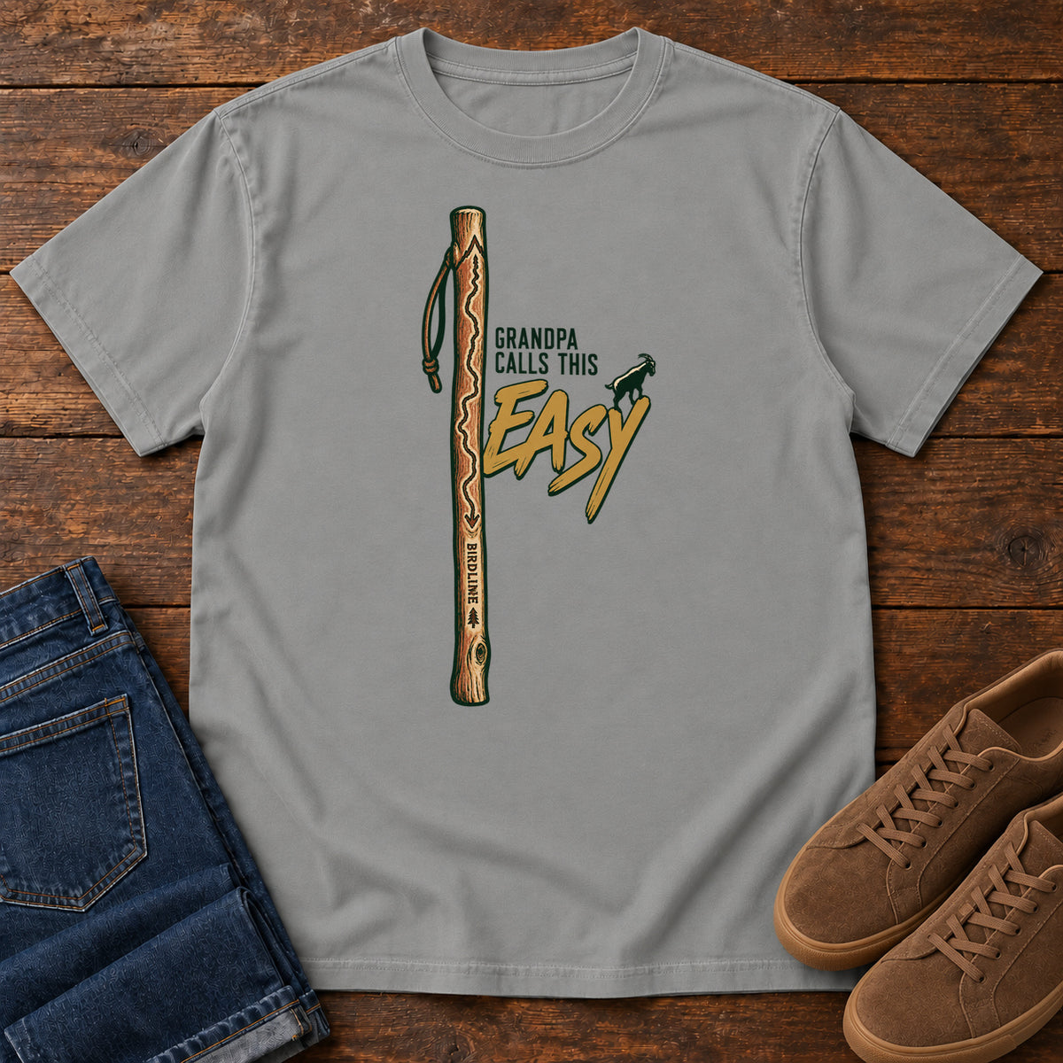 Grey Grandpa Calls This Easy T-Shirt with a wooden hiking stick beside green-and-gold lettering and a small goat silhouette; front flat lay on wood.