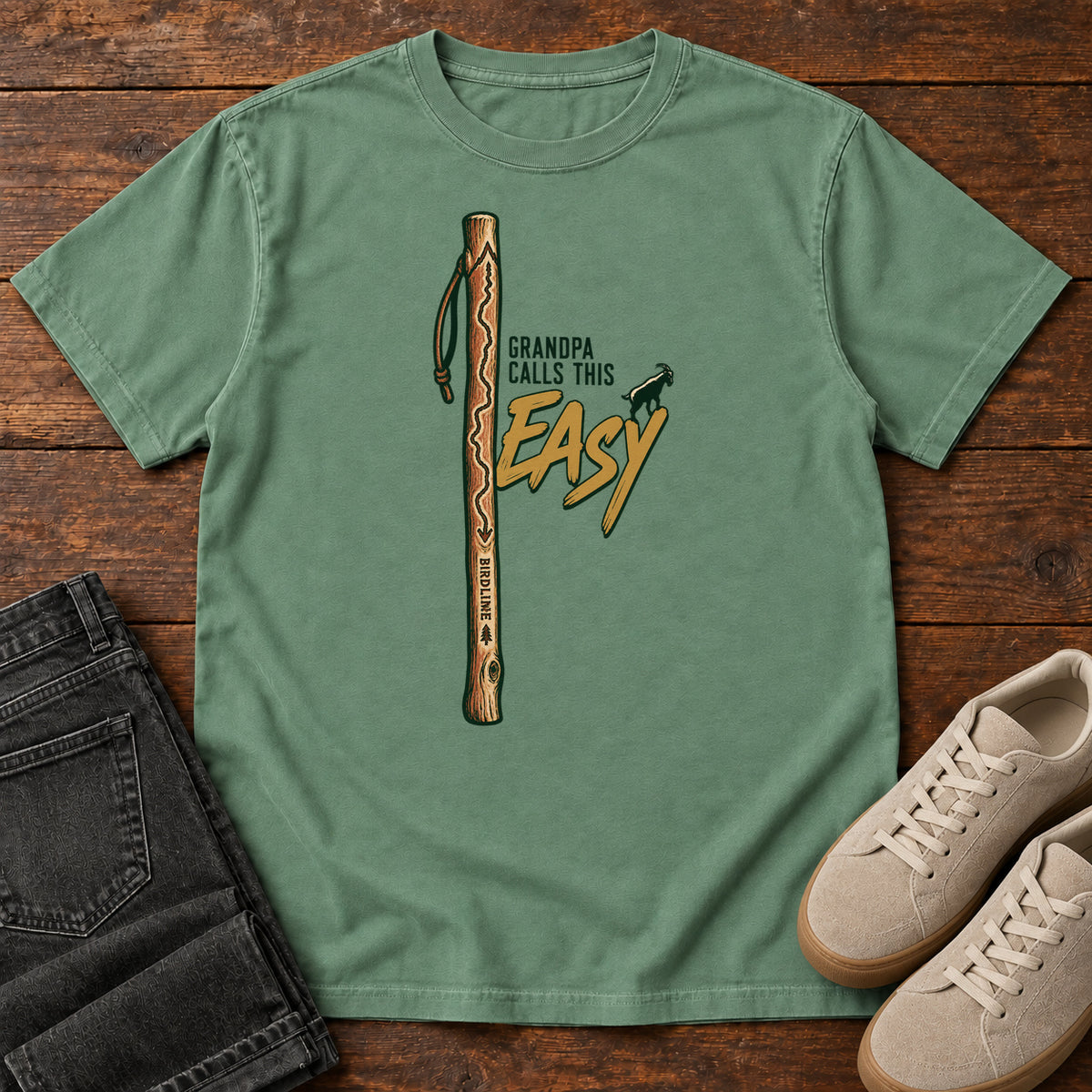 Light Green Grandpa Calls This Easy T-Shirt with a wooden hiking stick beside green-and-gold lettering and a small goat silhouette; front flat lay on wood.