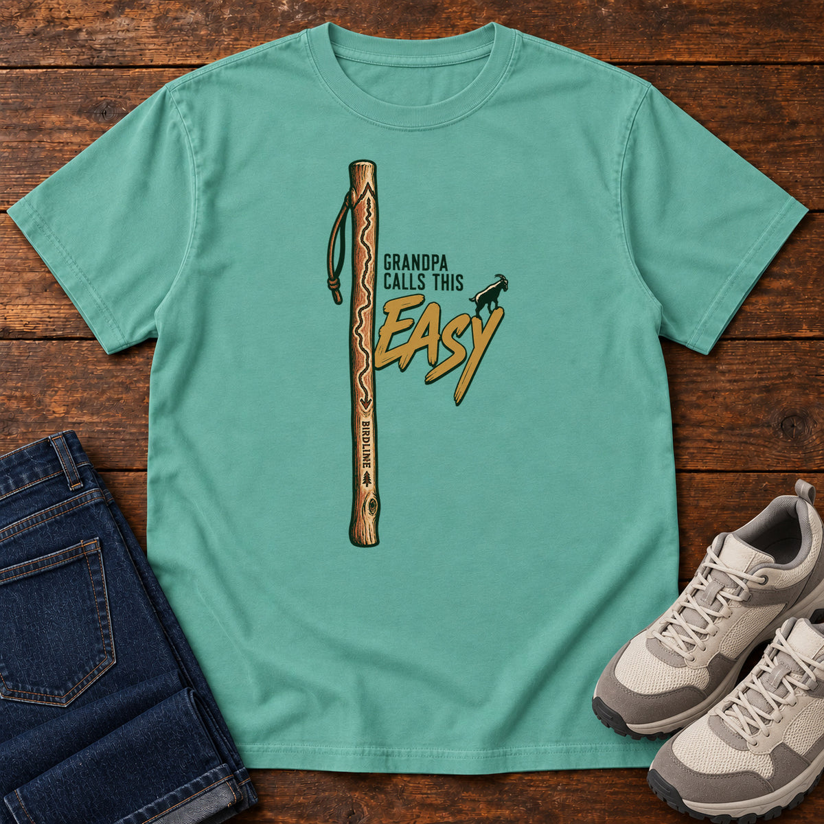 Seafoam Grandpa Calls This Easy T-Shirt with a wooden hiking stick beside green-and-gold lettering and a small goat silhouette; front flat lay on wood.