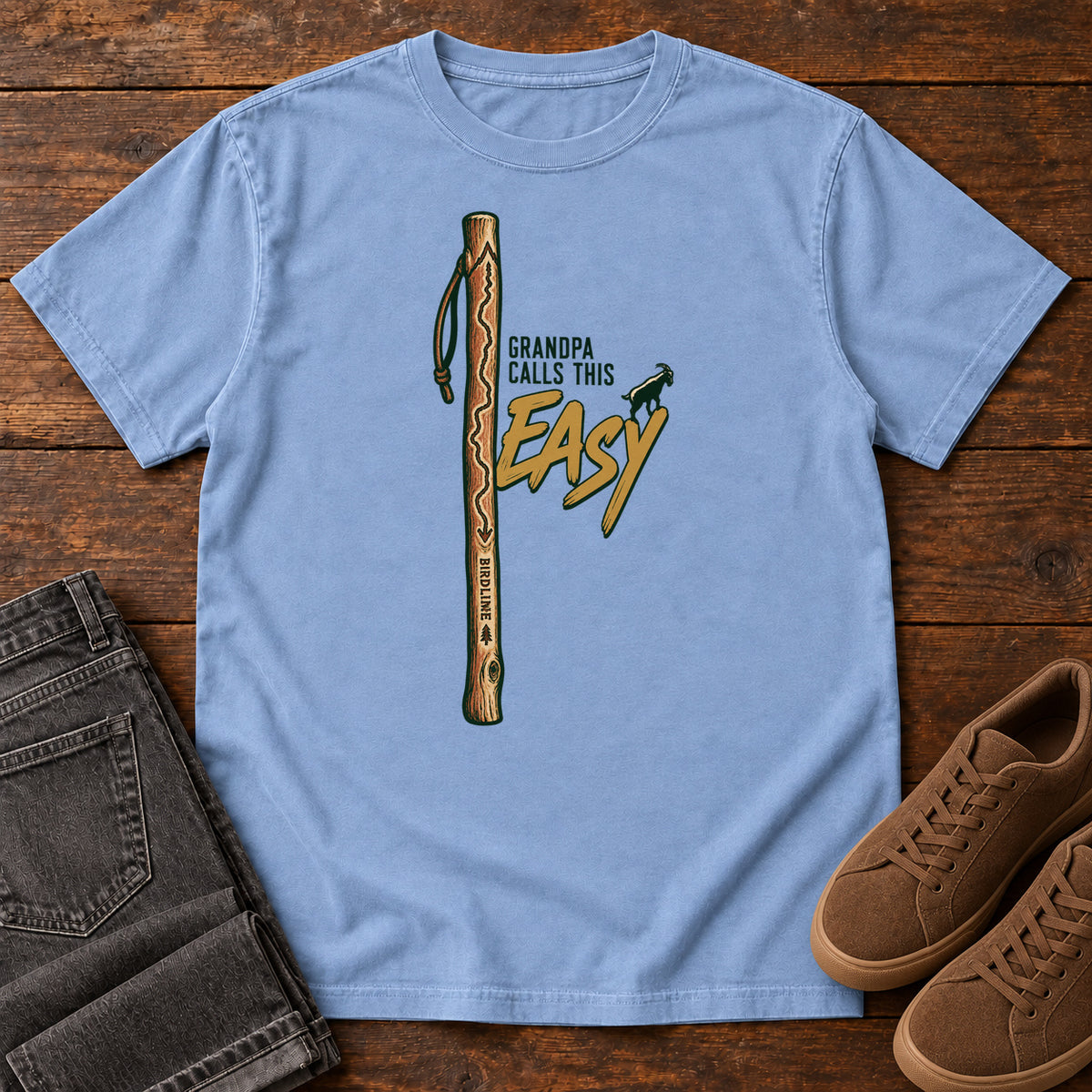 Washed Denim Grandpa Calls This Easy T-Shirt with a wooden hiking stick beside green-and-gold lettering and a small goat silhouette; front flat lay on wood.