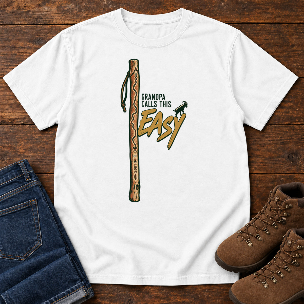 White Grandpa Calls This Easy T-Shirt with a wooden hiking stick beside green-and-gold lettering and a small goat silhouette; front flat lay on wood.