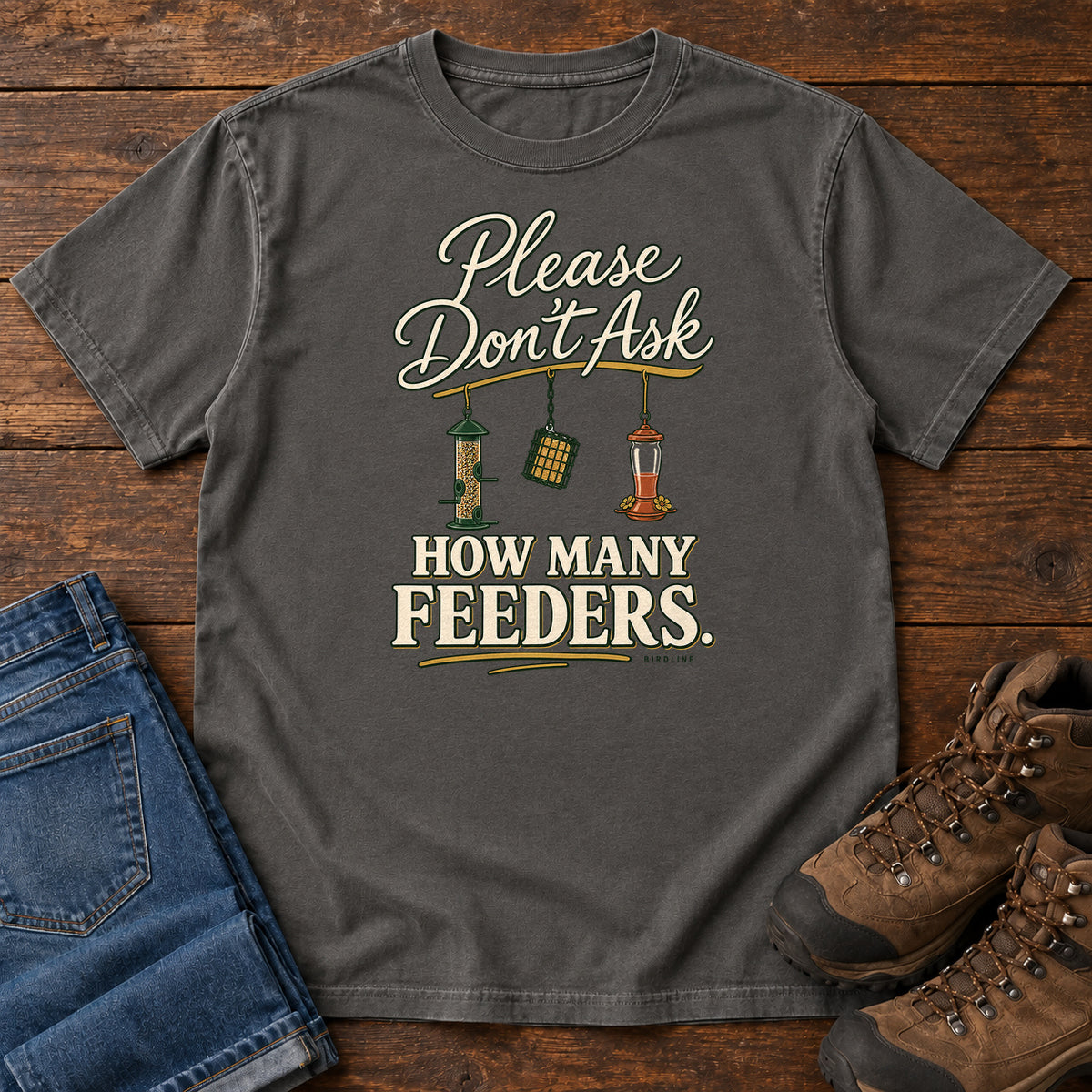 Pepper Please Don't Ask How Many Feeders T-Shirt with cream lettering and three hanging bird feeders, front flat lay on wood.