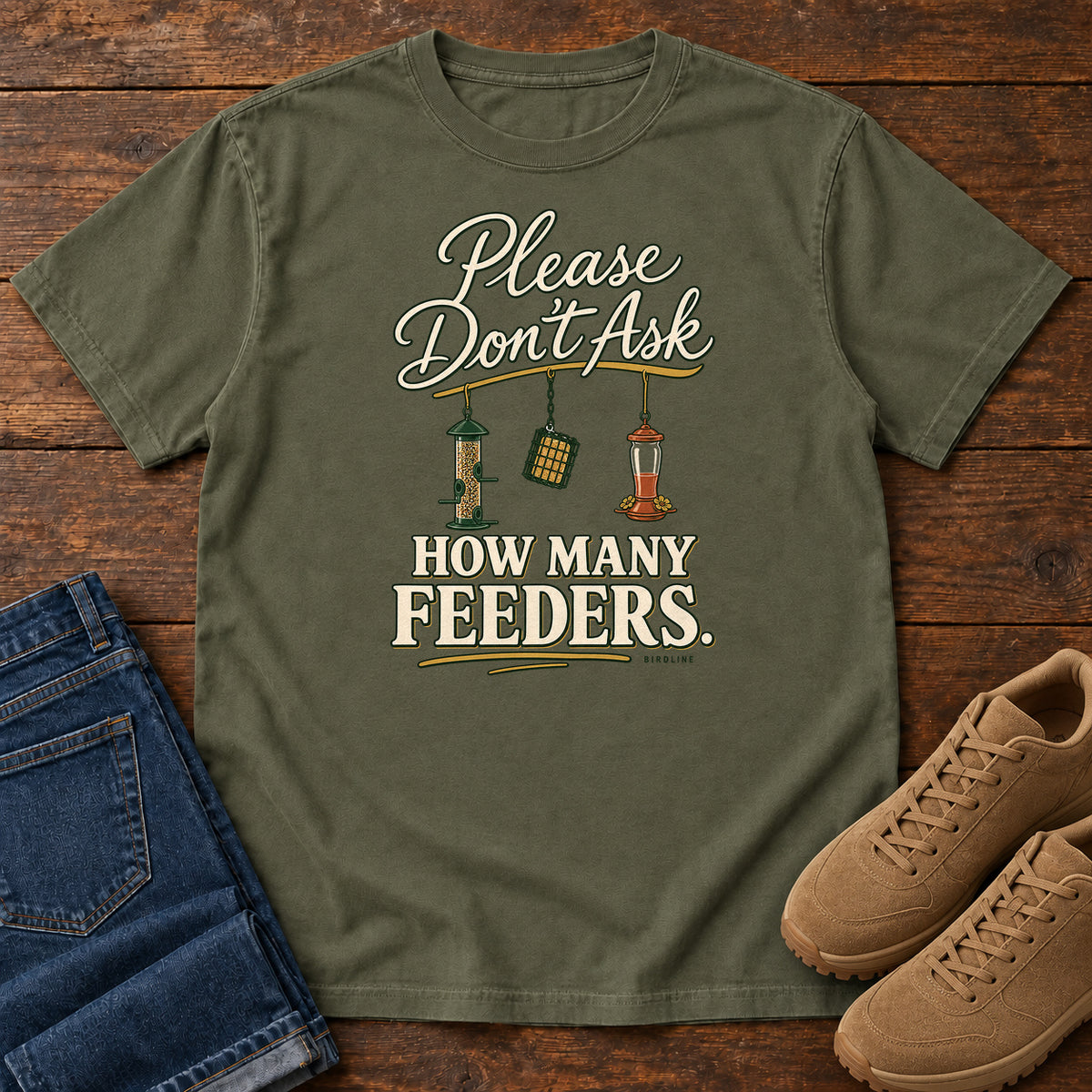 Sage Please Don't Ask How Many Feeders T-Shirt with cream lettering and three hanging bird feeders, front flat lay on wood.