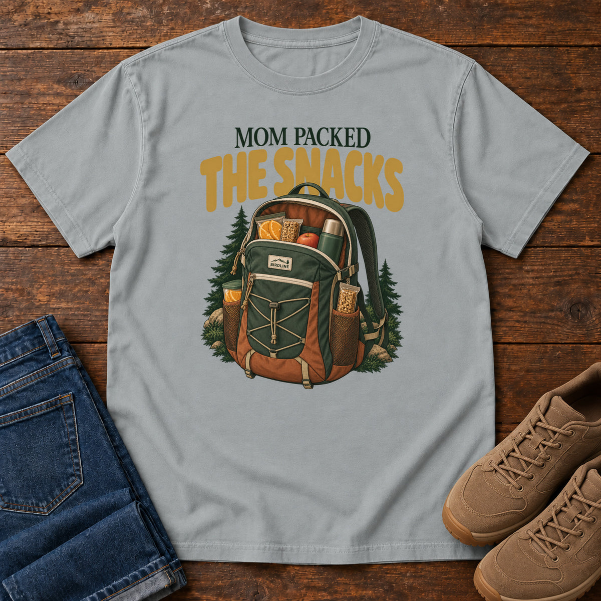 Granite Mom Packed the Snacks T-Shirt with a snack-filled green backpack framed by evergreens and gold lettering; front flat lay on wood.