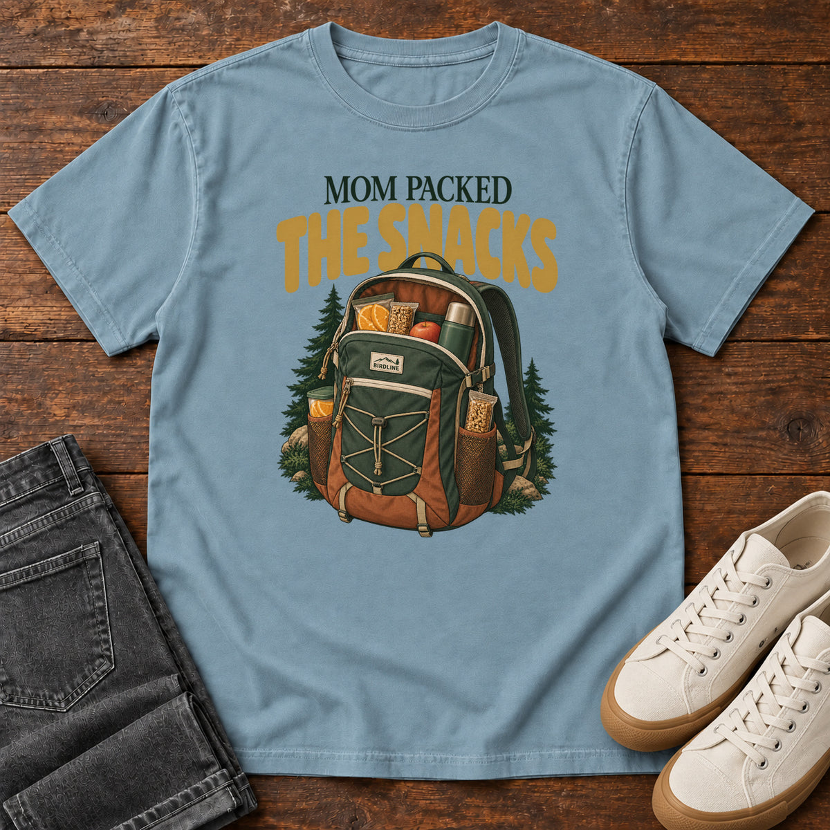 Ice Blue Mom Packed the Snacks T-Shirt with a snack-filled green backpack framed by evergreens and gold lettering; front flat lay on wood.