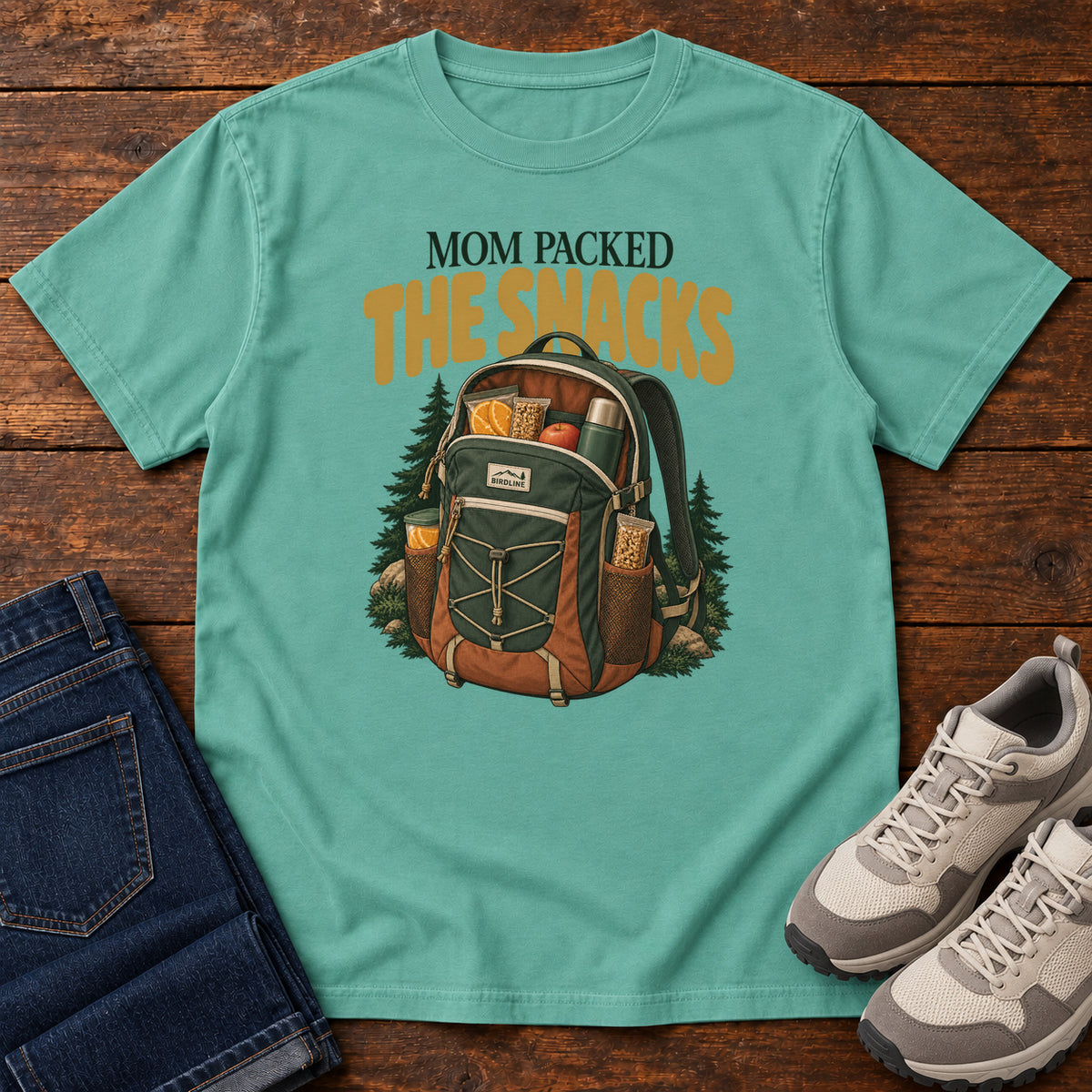 Seafoam Mom Packed the Snacks T-Shirt with a snack-filled green backpack framed by evergreens and gold lettering; front flat lay on wood.