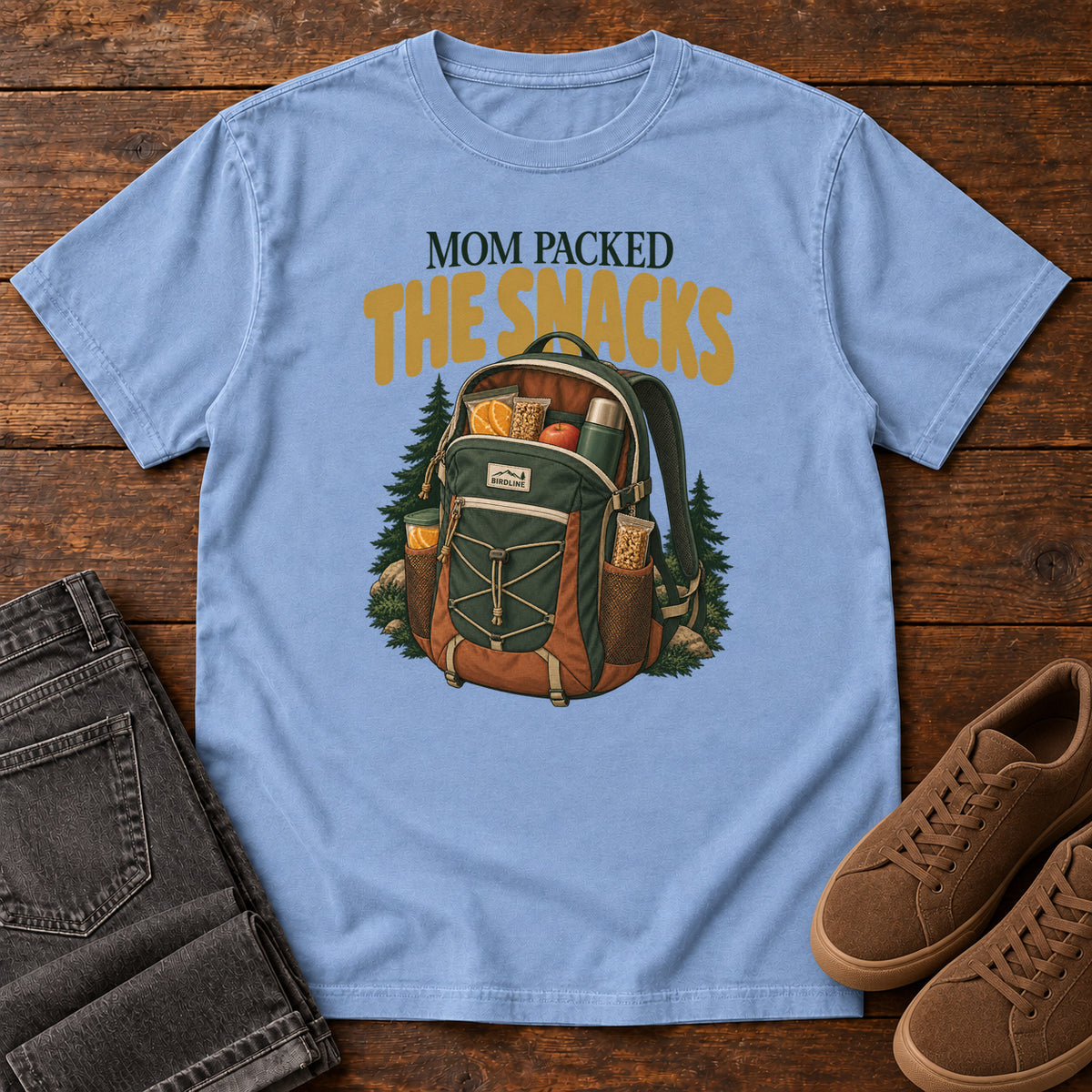 Washed Denim Mom Packed the Snacks T-Shirt with a snack-filled green backpack framed by evergreens and gold lettering; front flat lay on wood.