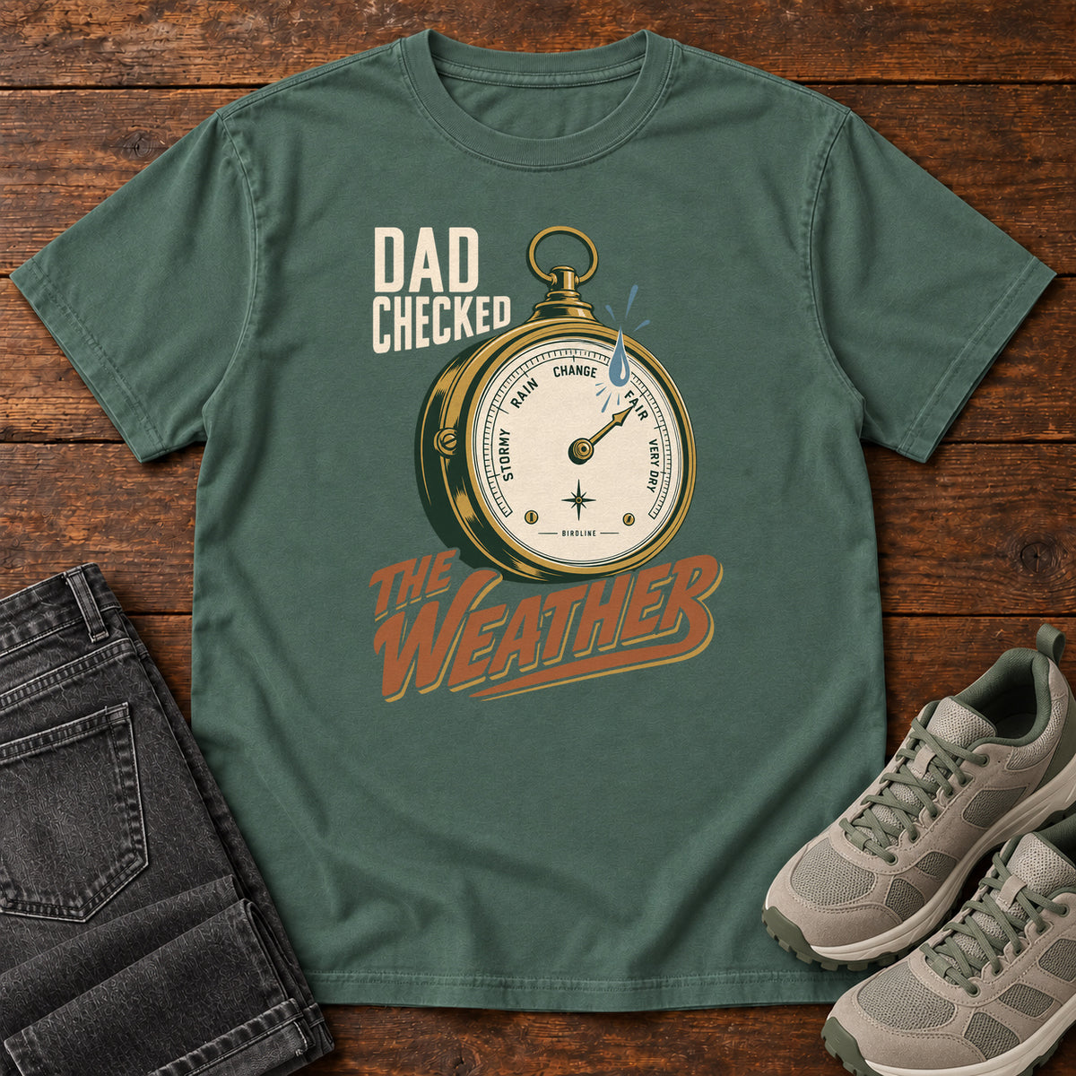 Blue Spruce Dad Checked the Weather T-shirt with a weather dial graphic and lettering, laid flat on a wooden surface.