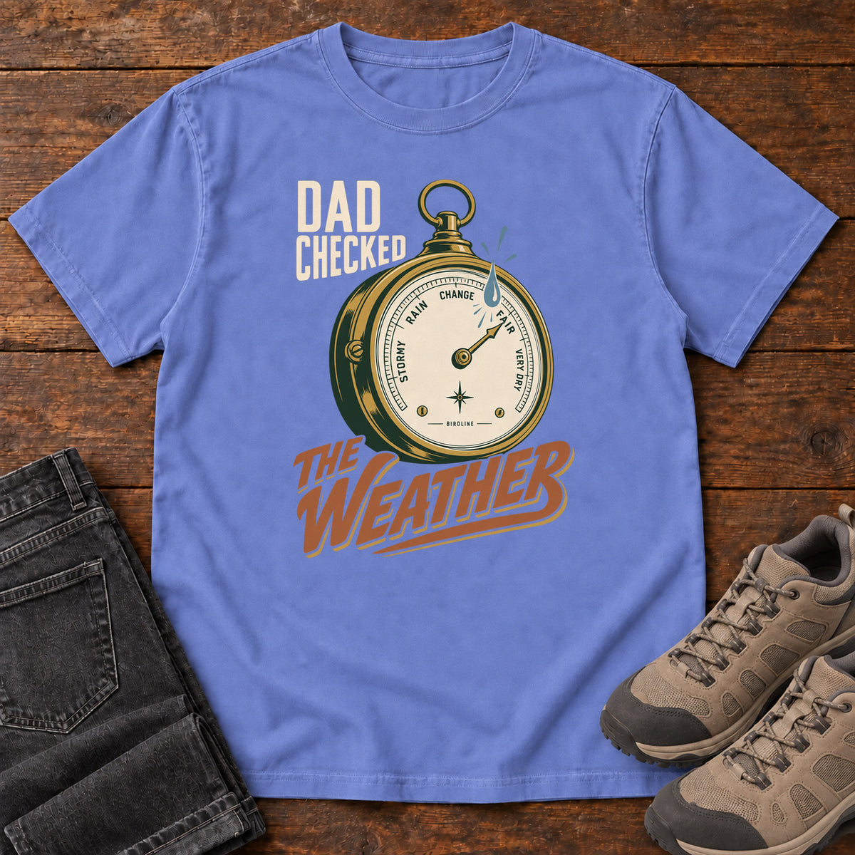 Flo Blue Dad Checked the Weather T-shirt with a weather dial graphic and lettering, laid flat on a wooden surface.