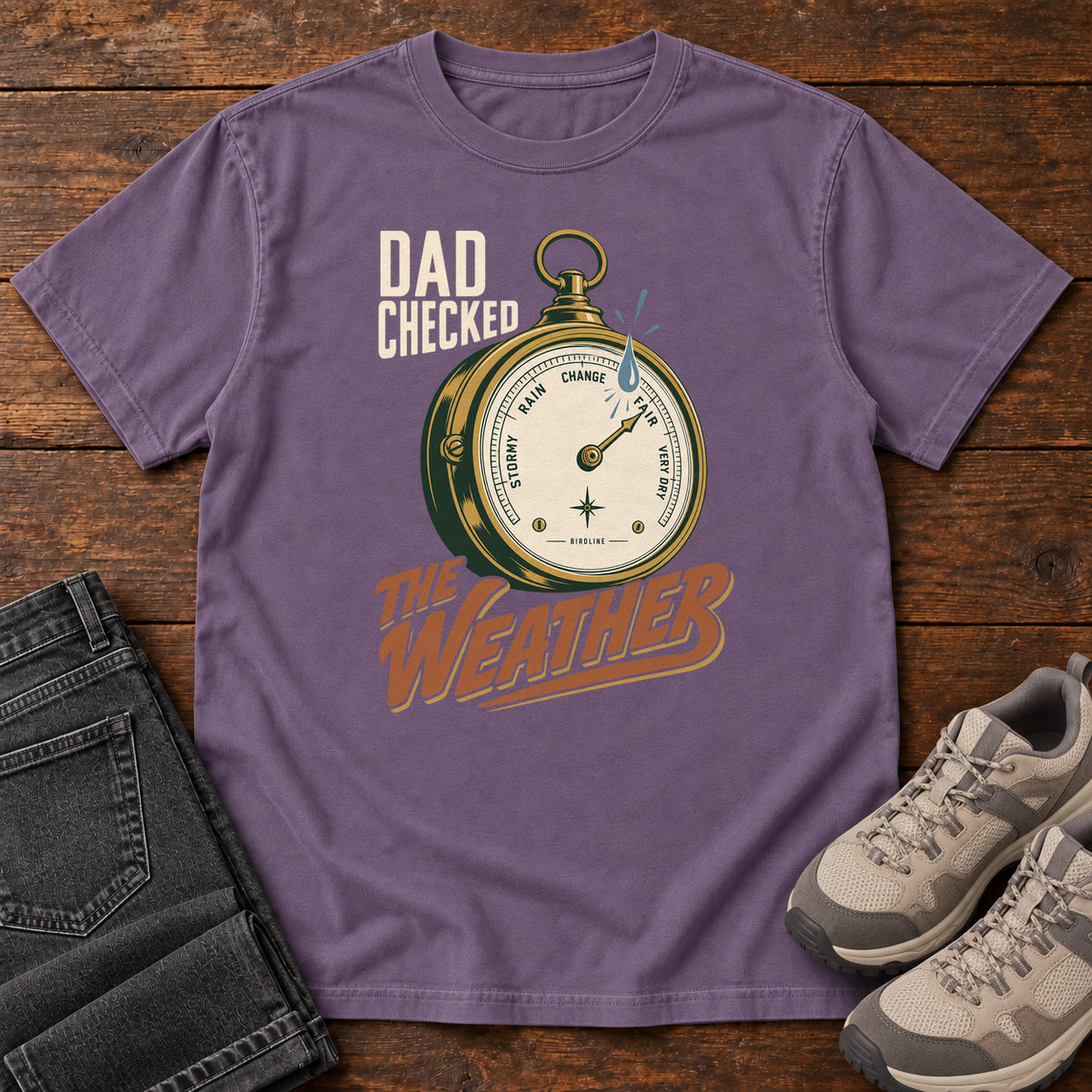 Grape Dad Checked the Weather T-shirt with a weather dial graphic and lettering, laid flat on a wooden surface.