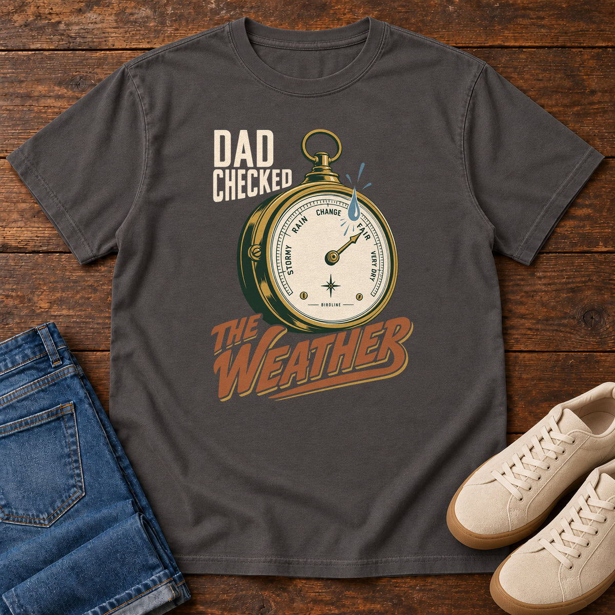Graphite Dad Checked the Weather T-shirt with a weather dial graphic and lettering, laid flat on a wooden surface.