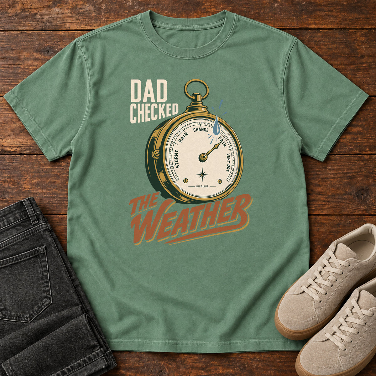 Light Green Dad Checked the Weather T-shirt with a weather dial graphic and lettering, laid flat on a wooden surface.