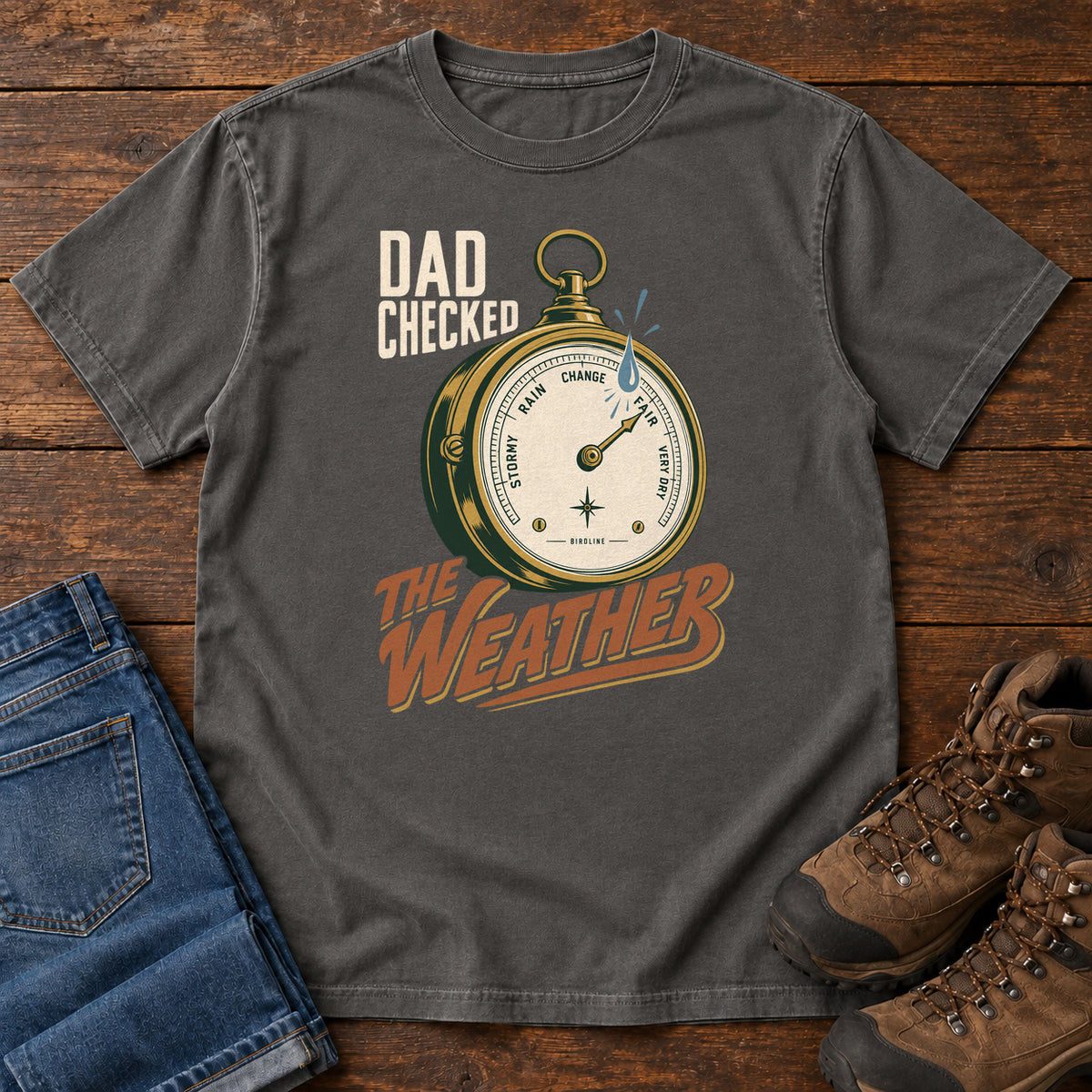 Pepper Dad Checked the Weather T-shirt with a weather dial graphic and lettering, laid flat on a wooden surface.