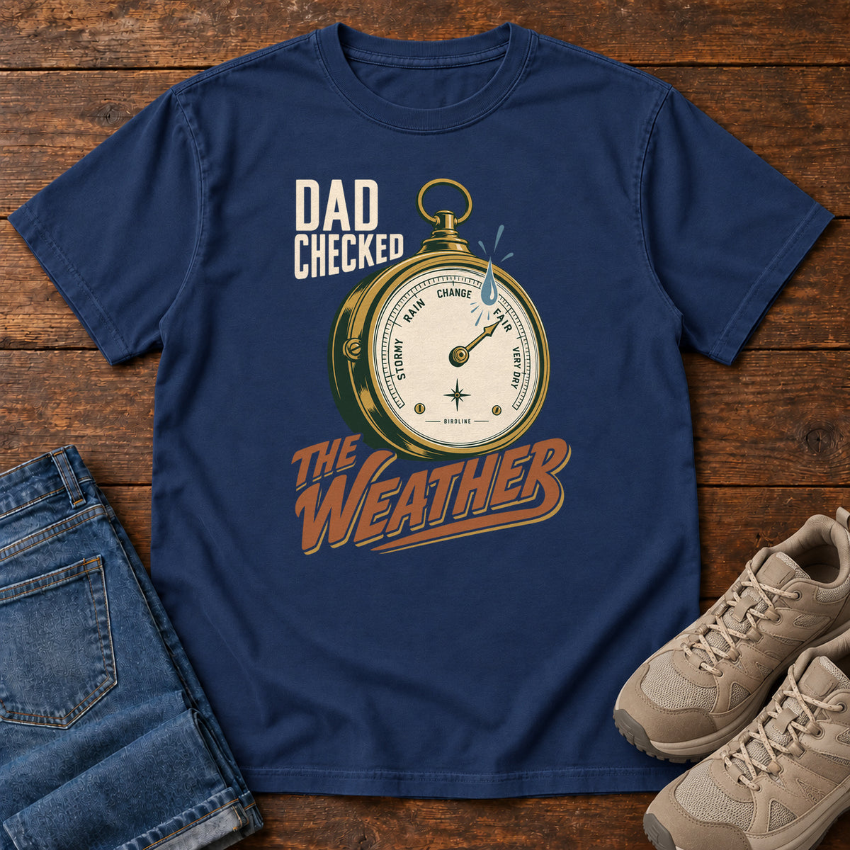 True Navy Dad Checked the Weather T-shirt with a weather dial graphic and lettering, laid flat on a wooden surface.