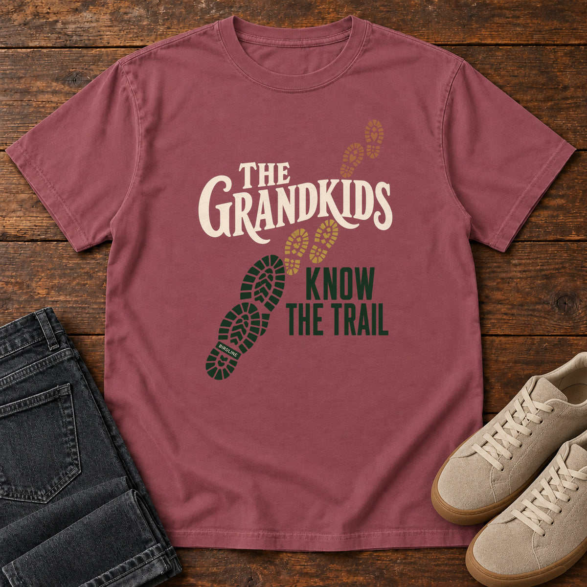 Brick The Grandkids Know the Trail T-Shirt with cream and green lettering beside a trail of green, gold and brown boot prints, front flat lay on wood.