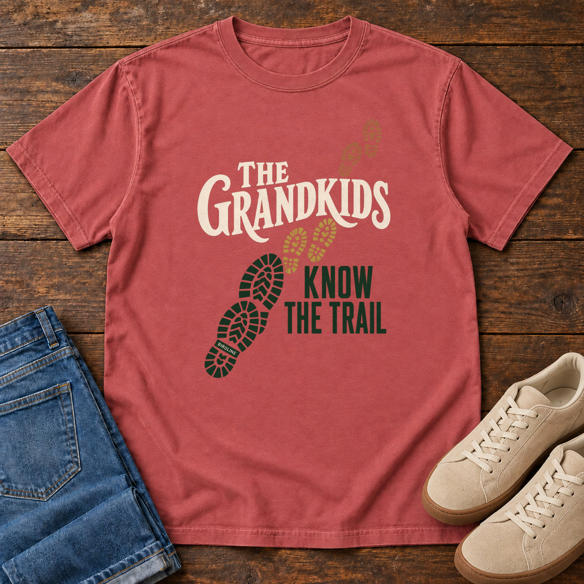 Crimson The Grandkids Know the Trail T-Shirt with cream and green lettering beside a trail of green, gold and brown boot prints, front flat lay on wood.