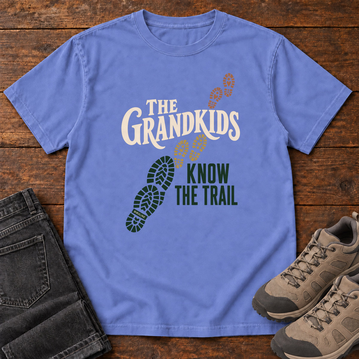 Flo Blue The Grandkids Know the Trail T-Shirt with cream and green lettering beside a trail of green, gold and brown boot prints, front flat lay on wood.