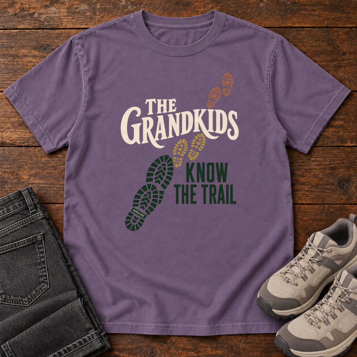 Grape The Grandkids Know the Trail T-Shirt with cream and green lettering beside a trail of green, gold and brown boot prints, front flat lay on wood.