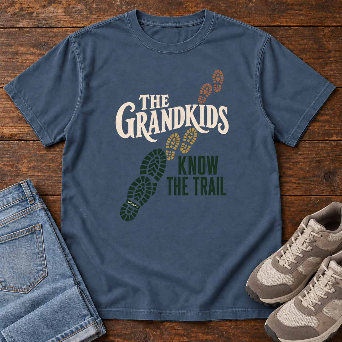 Midnight The Grandkids Know the Trail T-Shirt with cream and green lettering beside a trail of green, gold and brown boot prints, front flat lay on wood.