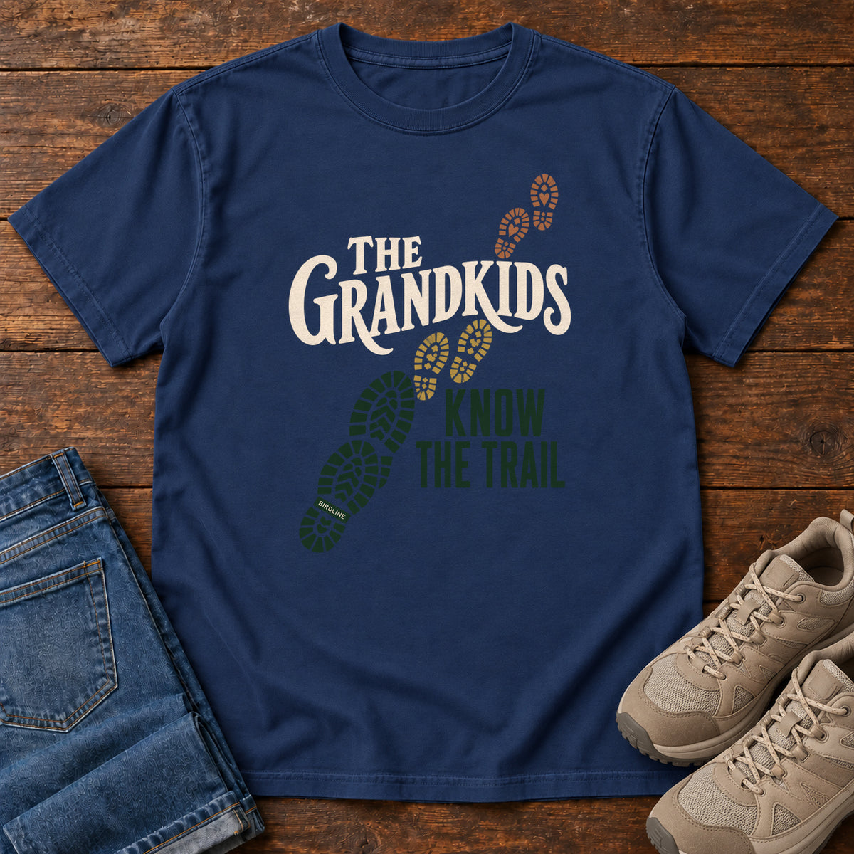 True Navy The Grandkids Know the Trail T-Shirt with cream and green lettering beside a trail of green, gold and brown boot prints, front flat lay on wood.