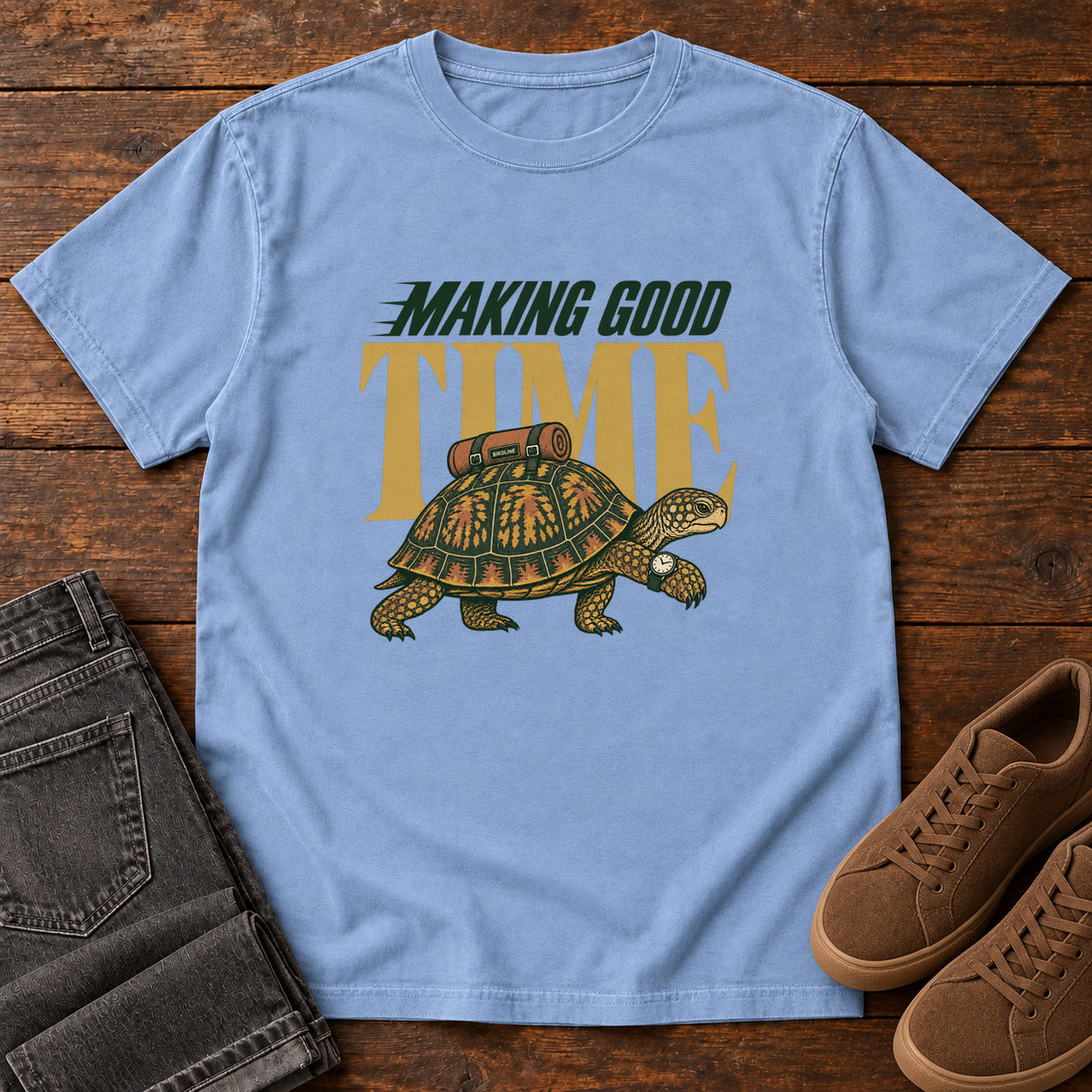 Washed Denim Making Good Time T-Shirt with a green-and-gold turtle carrying a bedroll beneath Making Good Time lettering, front flat lay on wood.