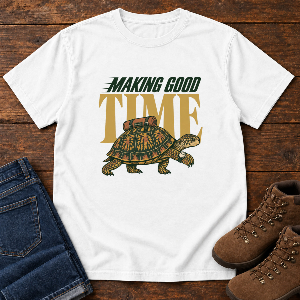 White Making Good Time T-Shirt with a green-and-gold turtle carrying a bedroll beneath Making Good Time lettering, front flat lay on wood.
