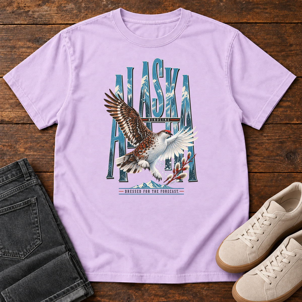 Orchid Alaska Dressed for the Forecast Eagle T-Shirt with a white-and-brown bird with raised wings over Alaska lettering and snowy mountains; front flat lay on wood.