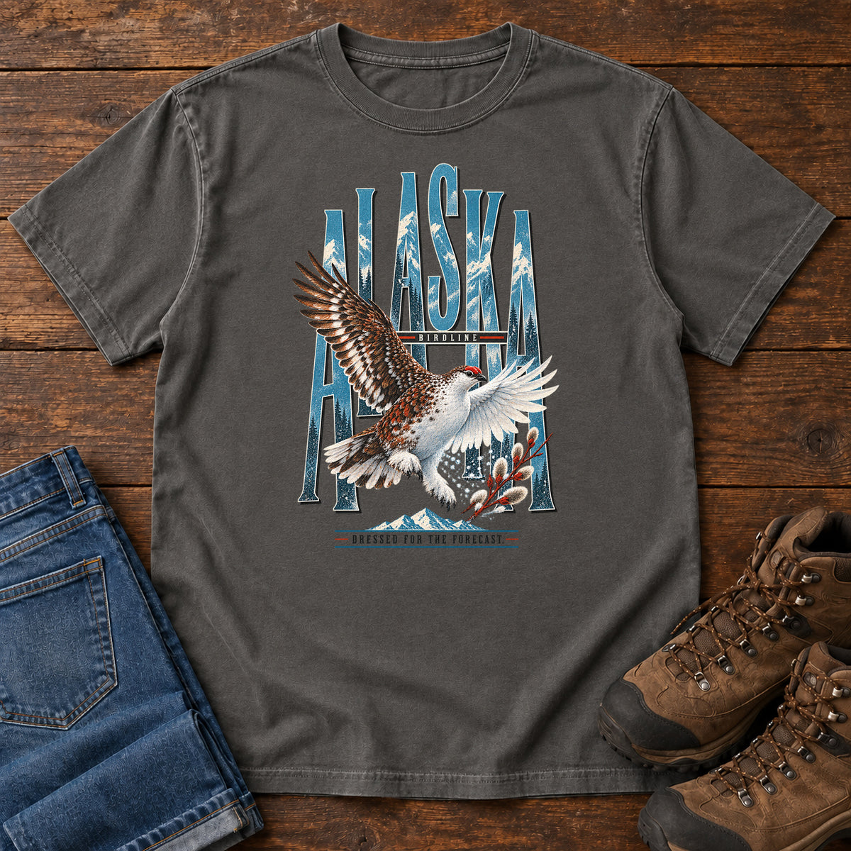 Pepper Alaska Dressed for the Forecast Eagle T-Shirt with a white-and-brown bird with raised wings over Alaska lettering and snowy mountains; front flat lay on wood.