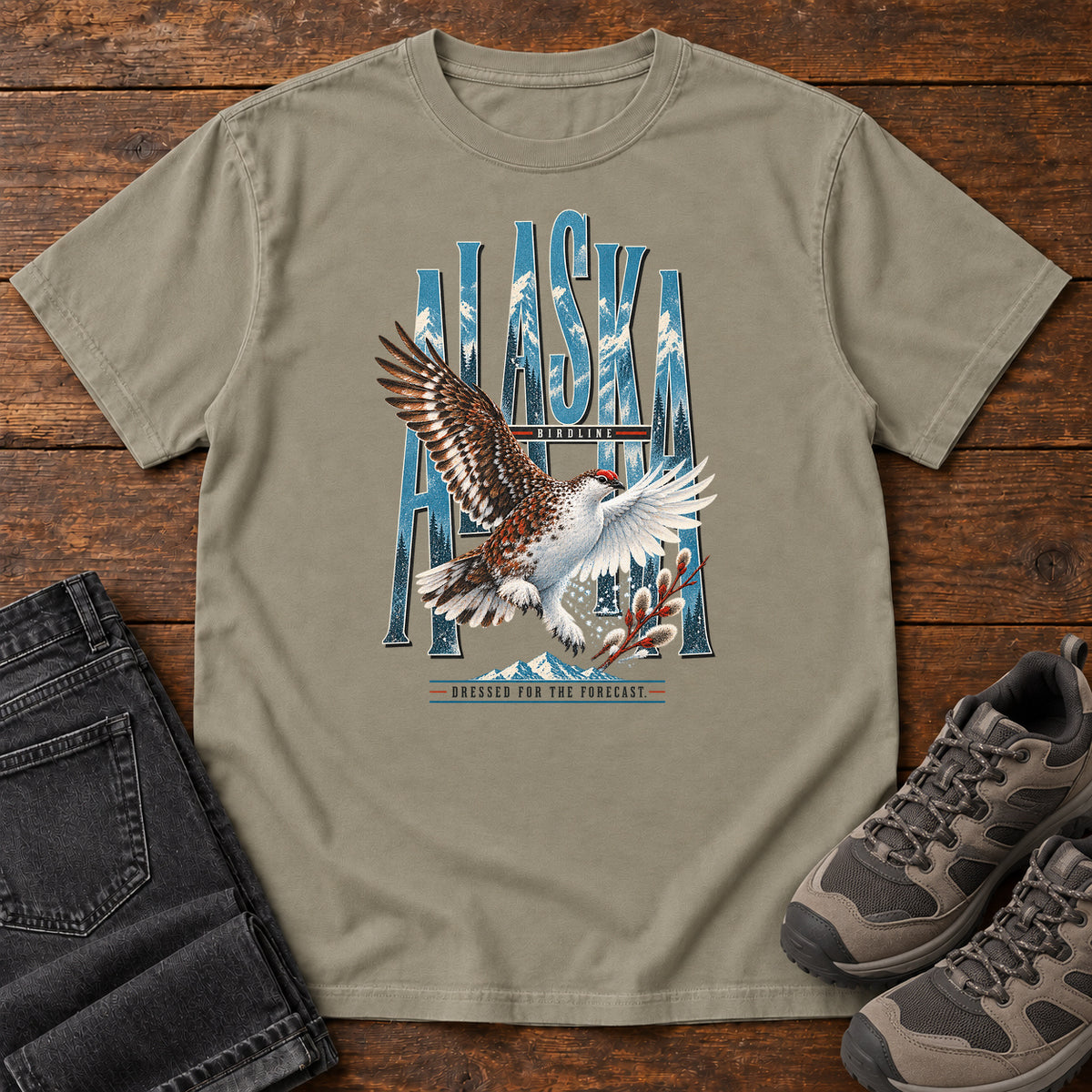 Sandstone Alaska Dressed for the Forecast Eagle T-Shirt with a white-and-brown bird with raised wings over Alaska lettering and snowy mountains; front flat lay on wood.