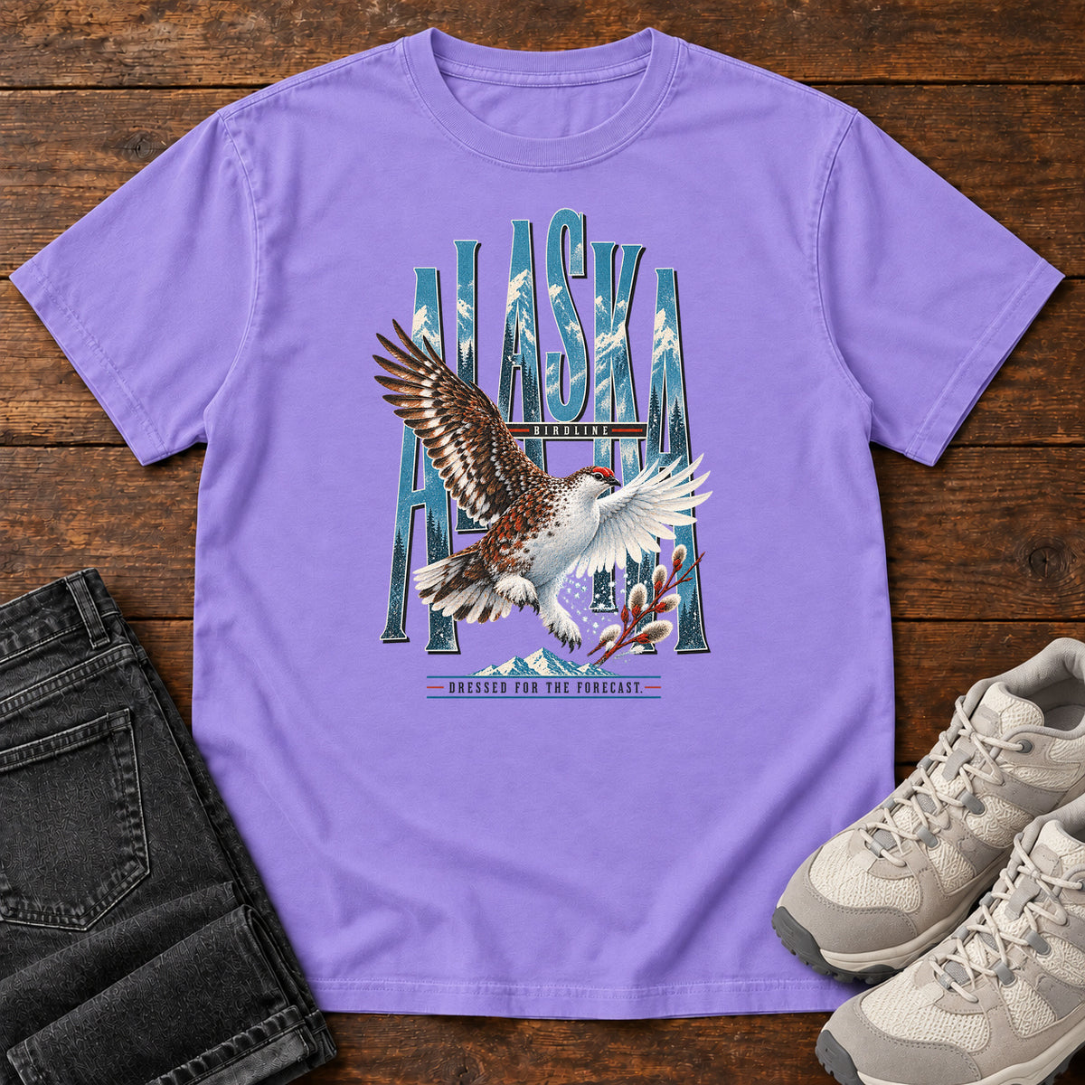 Violet Alaska Dressed for the Forecast Eagle T-Shirt with a white-and-brown bird with raised wings over Alaska lettering and snowy mountains; front flat lay on wood.