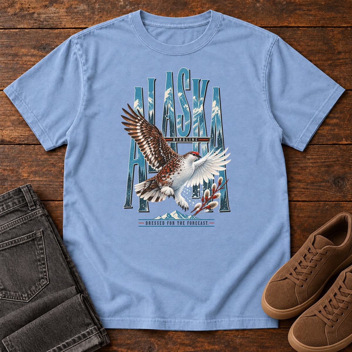 Washed Denim Alaska Dressed for the Forecast Eagle T-Shirt with a white-and-brown bird with raised wings over Alaska lettering and snowy mountains; front flat lay on wood.