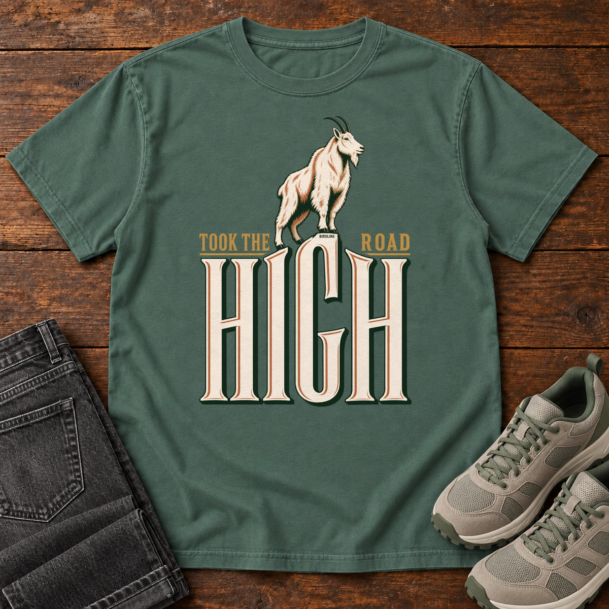 Took the High Road T-Shirt in Blue Spruce, front laid flat on wood, featuring a white mountain goat standing above oversized cream HIGH lettering.