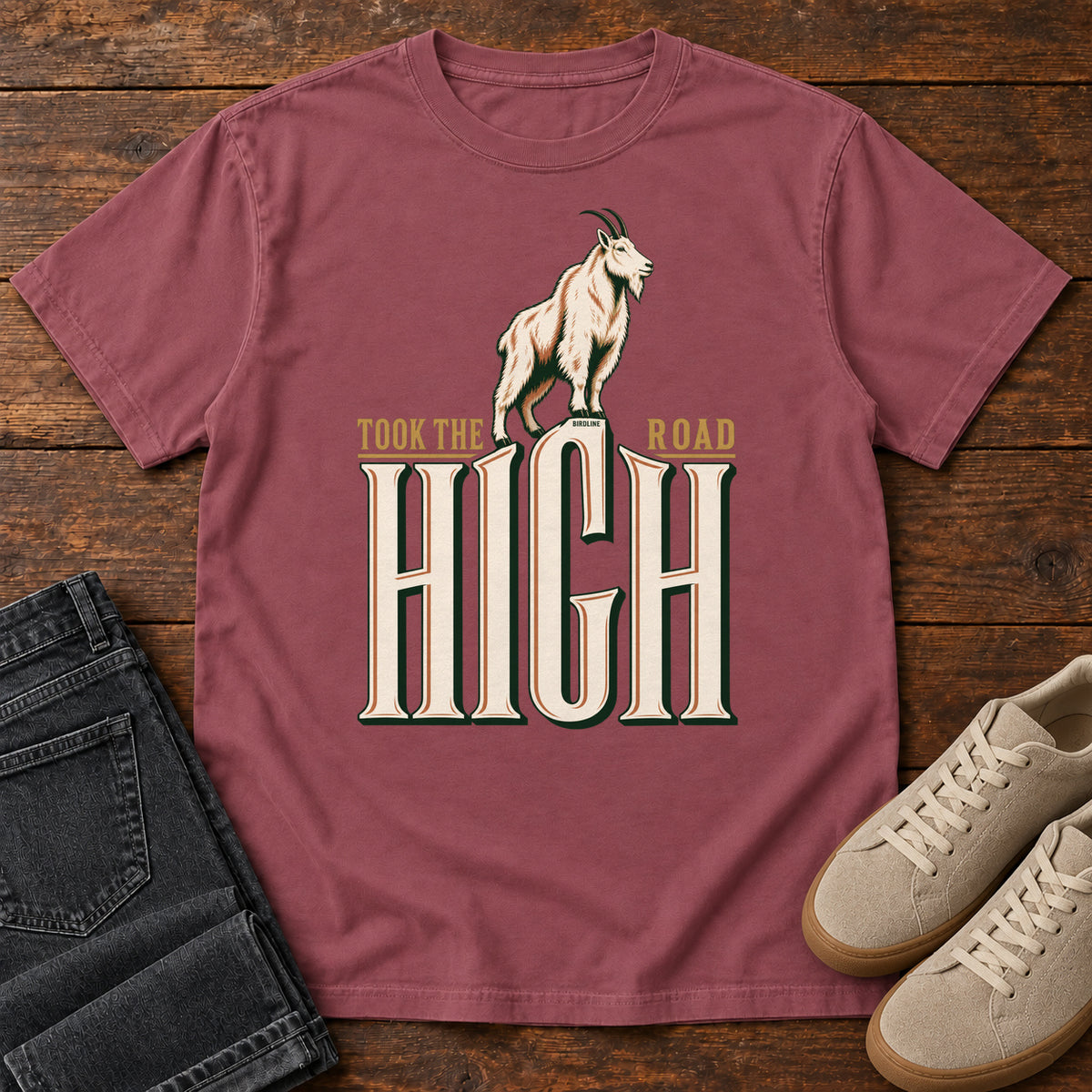 Took the High Road T-Shirt in Brick, front laid flat on wood, featuring a white mountain goat standing above oversized cream HIGH lettering.