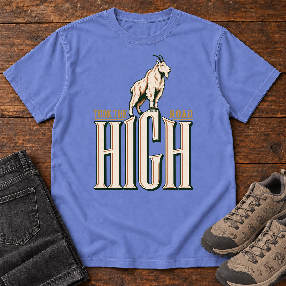 Took the High Road T-Shirt in Flo Blue, front laid flat on wood, featuring a white mountain goat standing above oversized cream HIGH lettering.