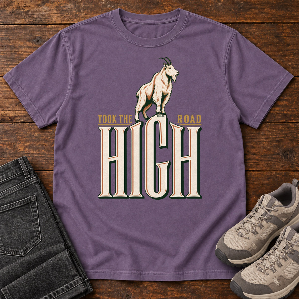 Took the High Road T-Shirt in Grape, front laid flat on wood, featuring a white mountain goat standing above oversized cream HIGH lettering.