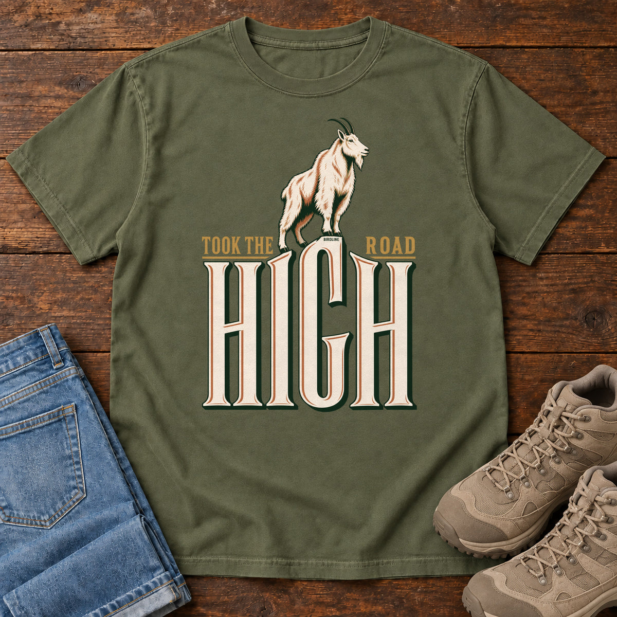 Took the High Road T-Shirt in Hemp, front laid flat on wood, featuring a white mountain goat standing above oversized cream HIGH lettering.