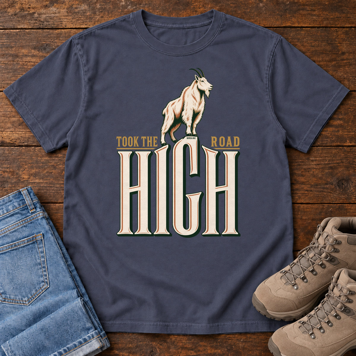 Took the High Road T-Shirt in Navy, front laid flat on wood, featuring a white mountain goat standing above oversized cream HIGH lettering.