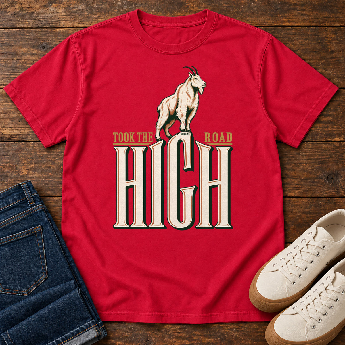 Took the High Road T-Shirt in Red, front laid flat on wood, featuring a white mountain goat standing above oversized cream HIGH lettering.