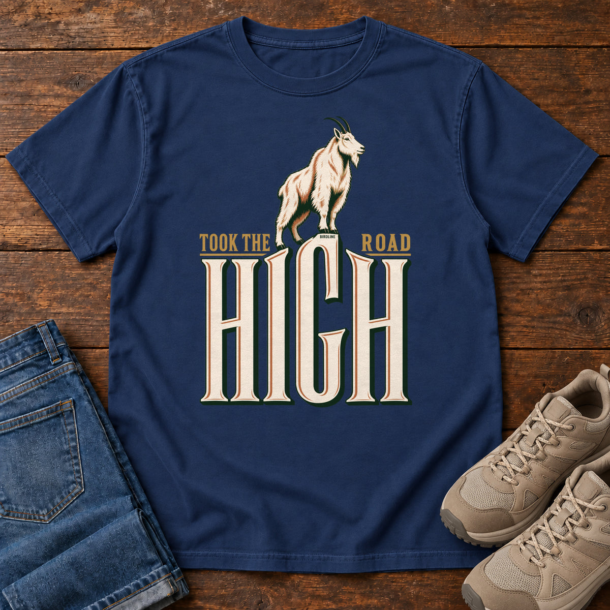 Took the High Road T-Shirt in True Navy, front laid flat on wood, featuring a white mountain goat standing above oversized cream HIGH lettering.