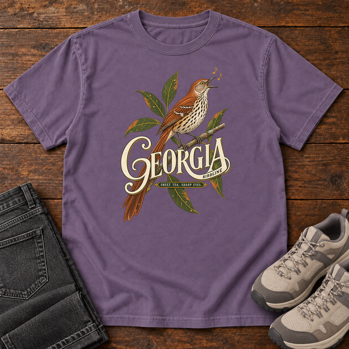 Grape Georgia Sharp Eyes T-Shirt with a brown, speckle-breasted bird on a leafy branch above Georgia lettering, front flat lay on wood.