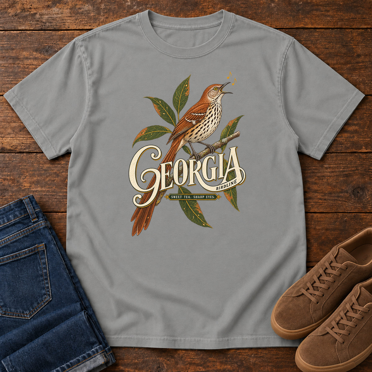 Grey Georgia Sharp Eyes T-Shirt with a brown, speckle-breasted bird on a leafy branch above Georgia lettering, front flat lay on wood.