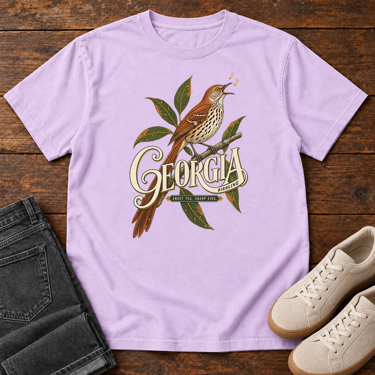 Orchid Georgia Sharp Eyes T-Shirt with a brown, speckle-breasted bird on a leafy branch above Georgia lettering, front flat lay on wood.
