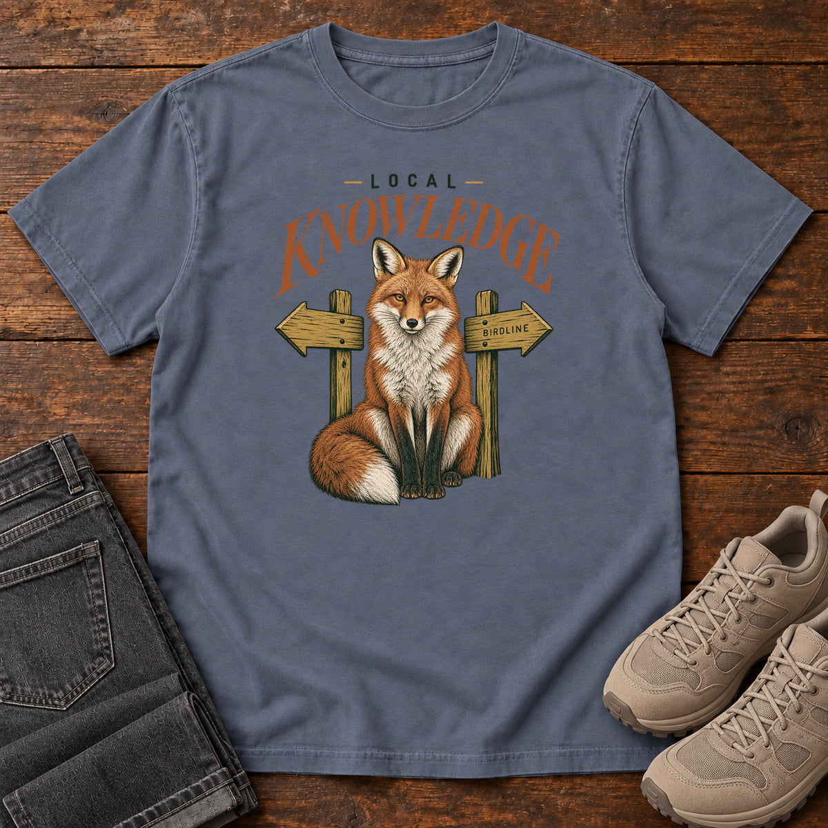 Denim Local Knowledge T-Shirt with a seated fox between wooden direction signs beneath Local Knowledge lettering; front flat lay on wood.