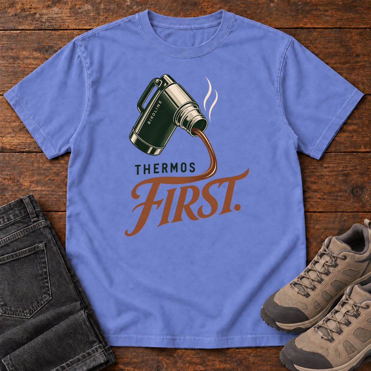 Flo Blue Thermos First T-Shirt with a green thermos pouring a steaming drink into rust-colored script, front flat lay on wood.