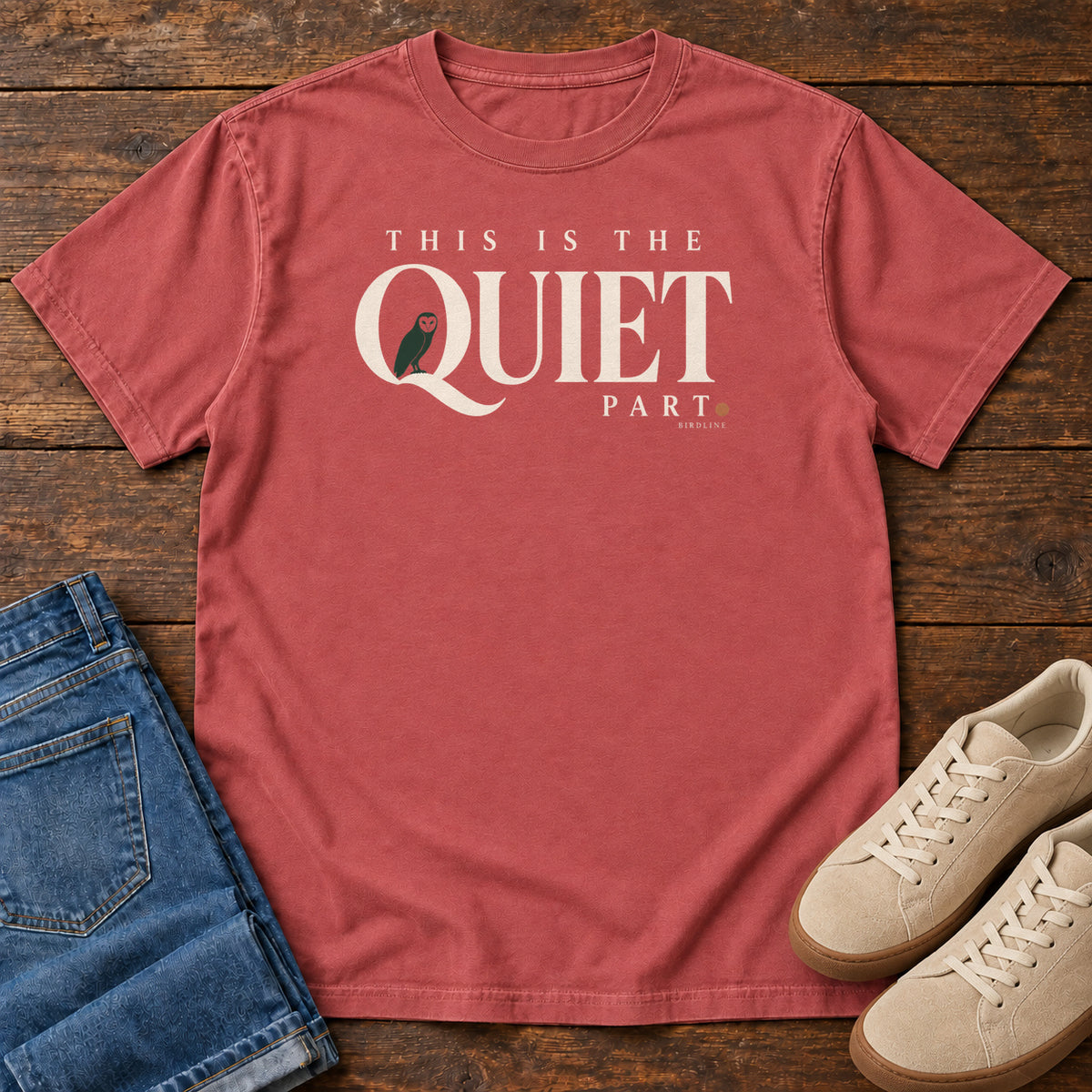 Crimson This Is the Quiet Part T-Shirt with cream This Is the Quiet Part lettering with a small green bird inside the Q, front flat lay on wood.