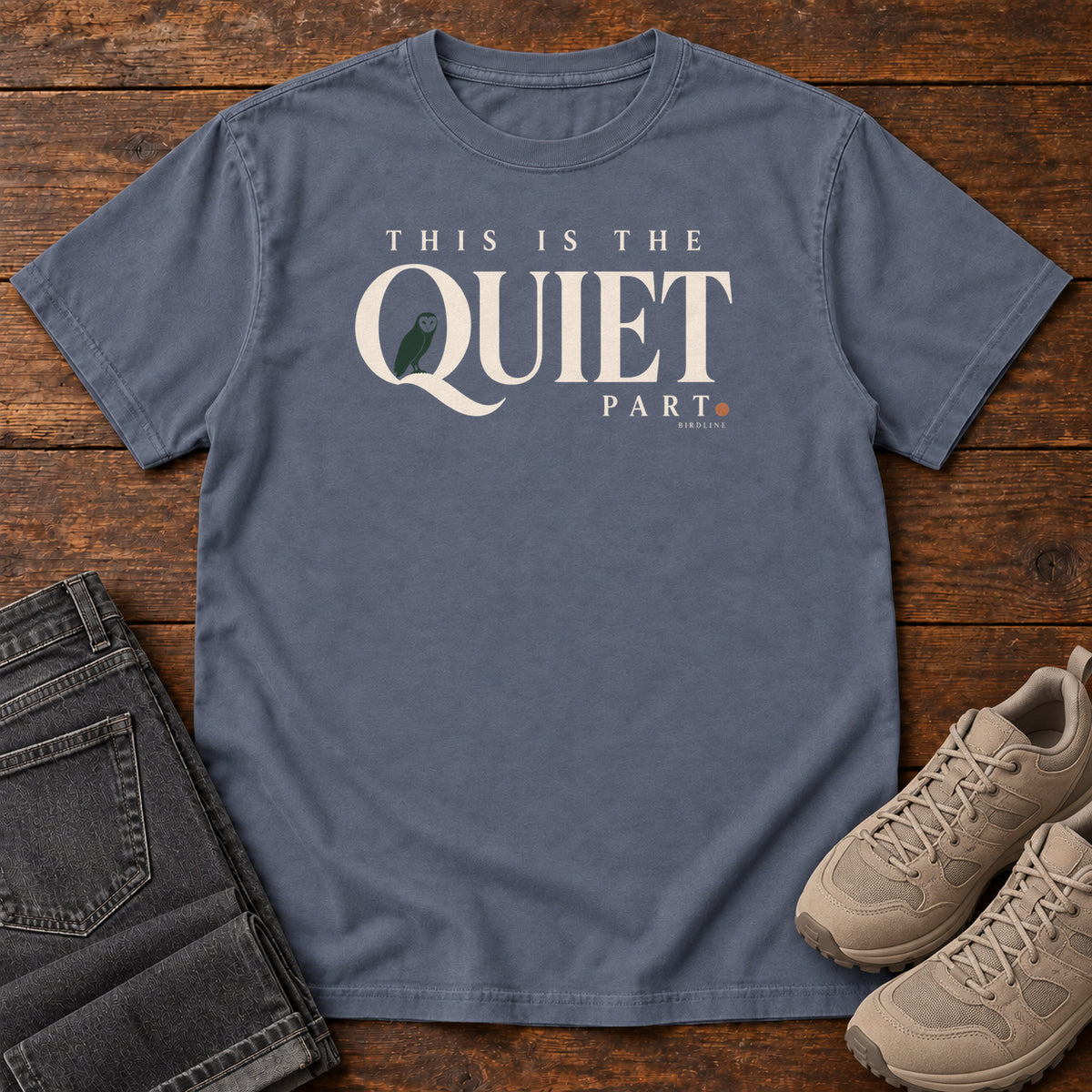 Denim This Is the Quiet Part T-Shirt with cream This Is the Quiet Part lettering with a small green bird inside the Q, front flat lay on wood.