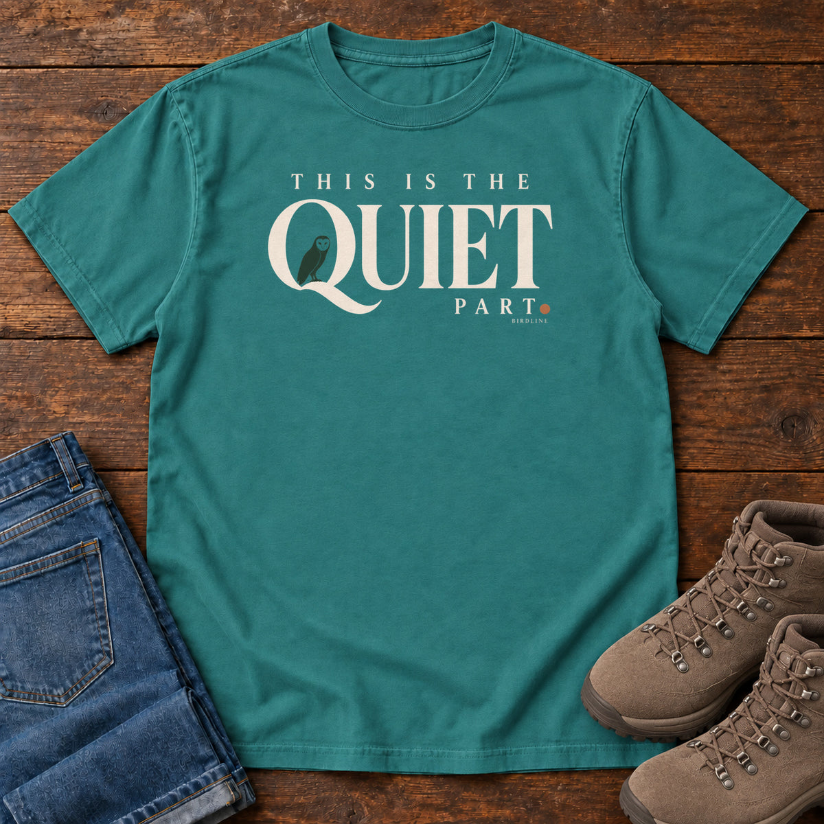 Emerald This Is the Quiet Part T-Shirt with cream This Is the Quiet Part lettering with a small green bird inside the Q, front flat lay on wood.