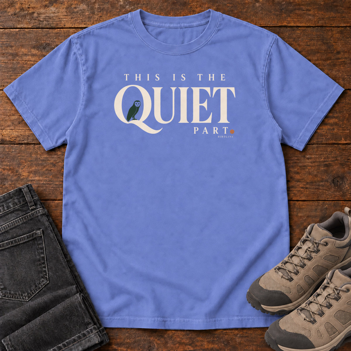 Flo Blue This Is the Quiet Part T-Shirt with cream This Is the Quiet Part lettering with a small green bird inside the Q, front flat lay on wood.