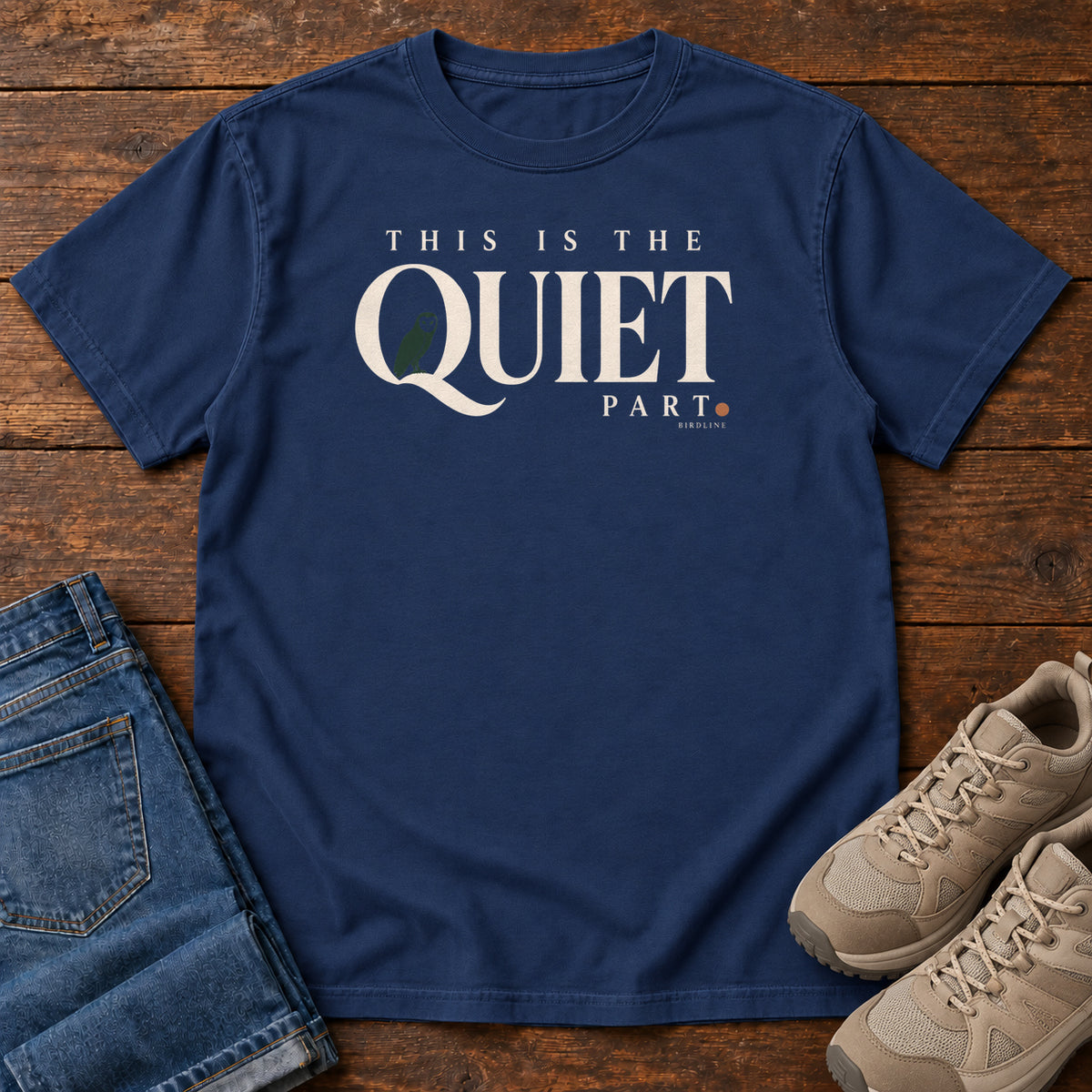True Navy This Is the Quiet Part T-Shirt with cream This Is the Quiet Part lettering with a small green bird inside the Q, front flat lay on wood.