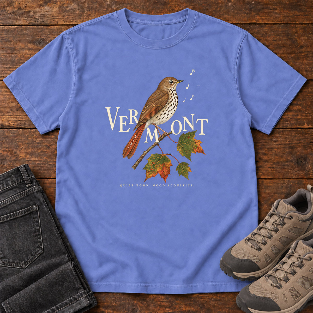 Flo Blue Vermont Quiet Town Good Acoustics Thrush T-Shirt with a speckled brown bird, autumn leaves and musical notes; front flat lay on wood.